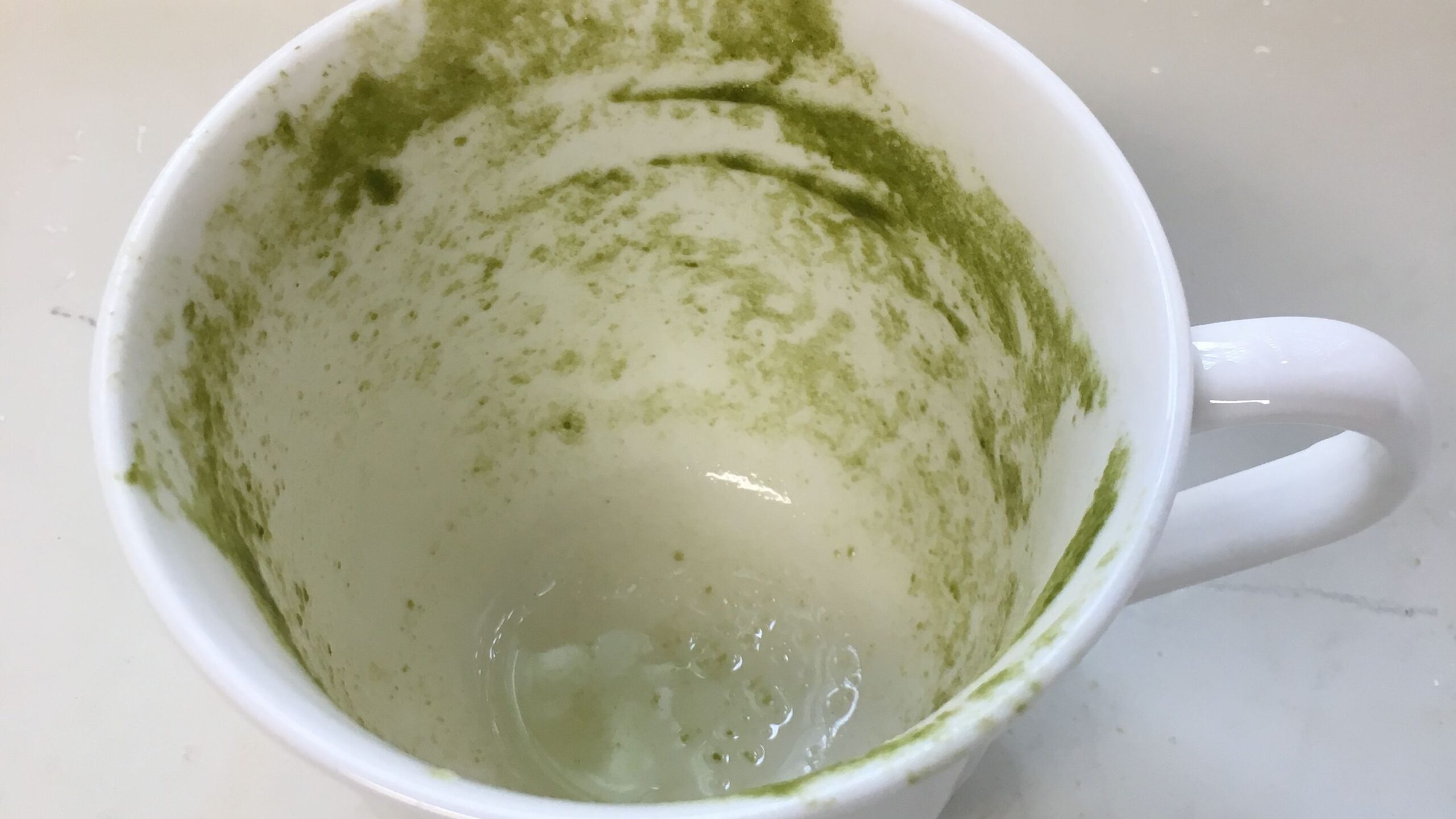 Empty white mug with green stains on the inside, possibly from matcha or a similar beverage, on a light-colored surface.