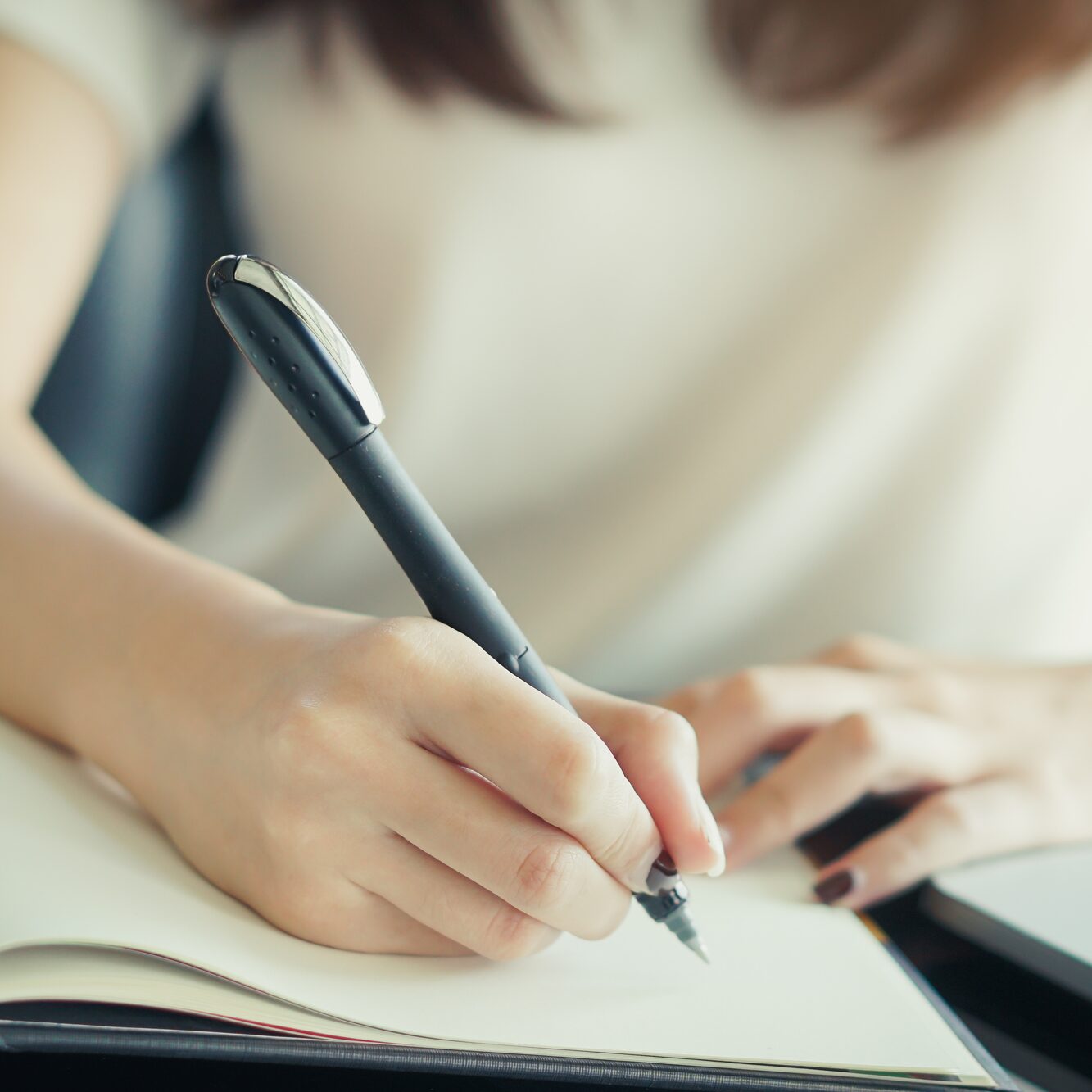 Depositphotos_642219732_L Person writing in a notebook with a black pen, wearing a light-colored shirt.