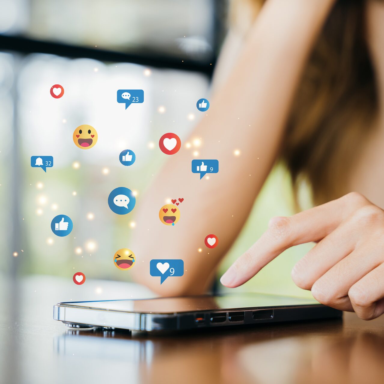 A hand interacts with a smartphone on a table. Above the phone, social media reaction icons, like hearts and thumbs up, are floating.