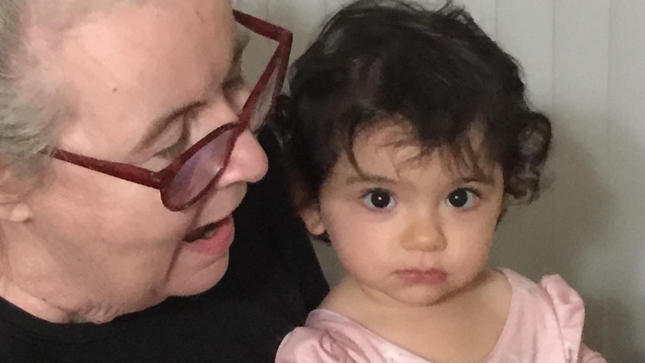 An older woman with glasses holds a young child wearing a pink dress; the woman is looking at the child.