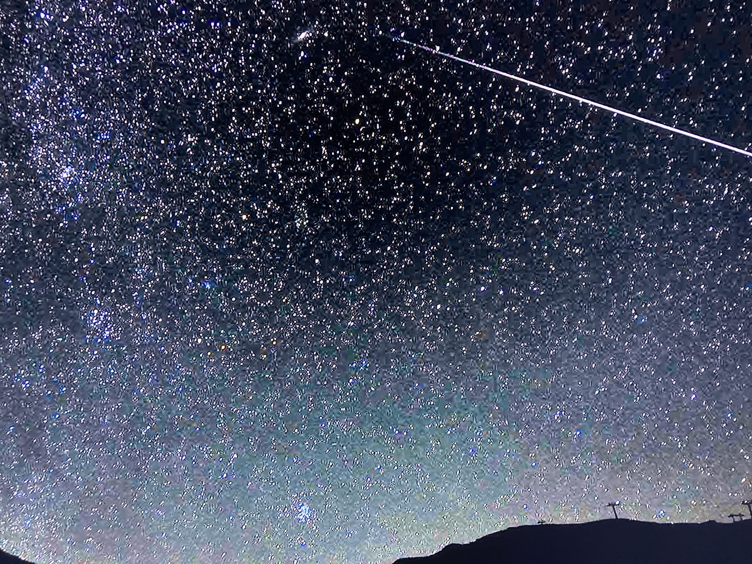 Night sky filled with stars, a bright meteor streaks across, and a dark silhouette of mountains is visible at the bottom.
