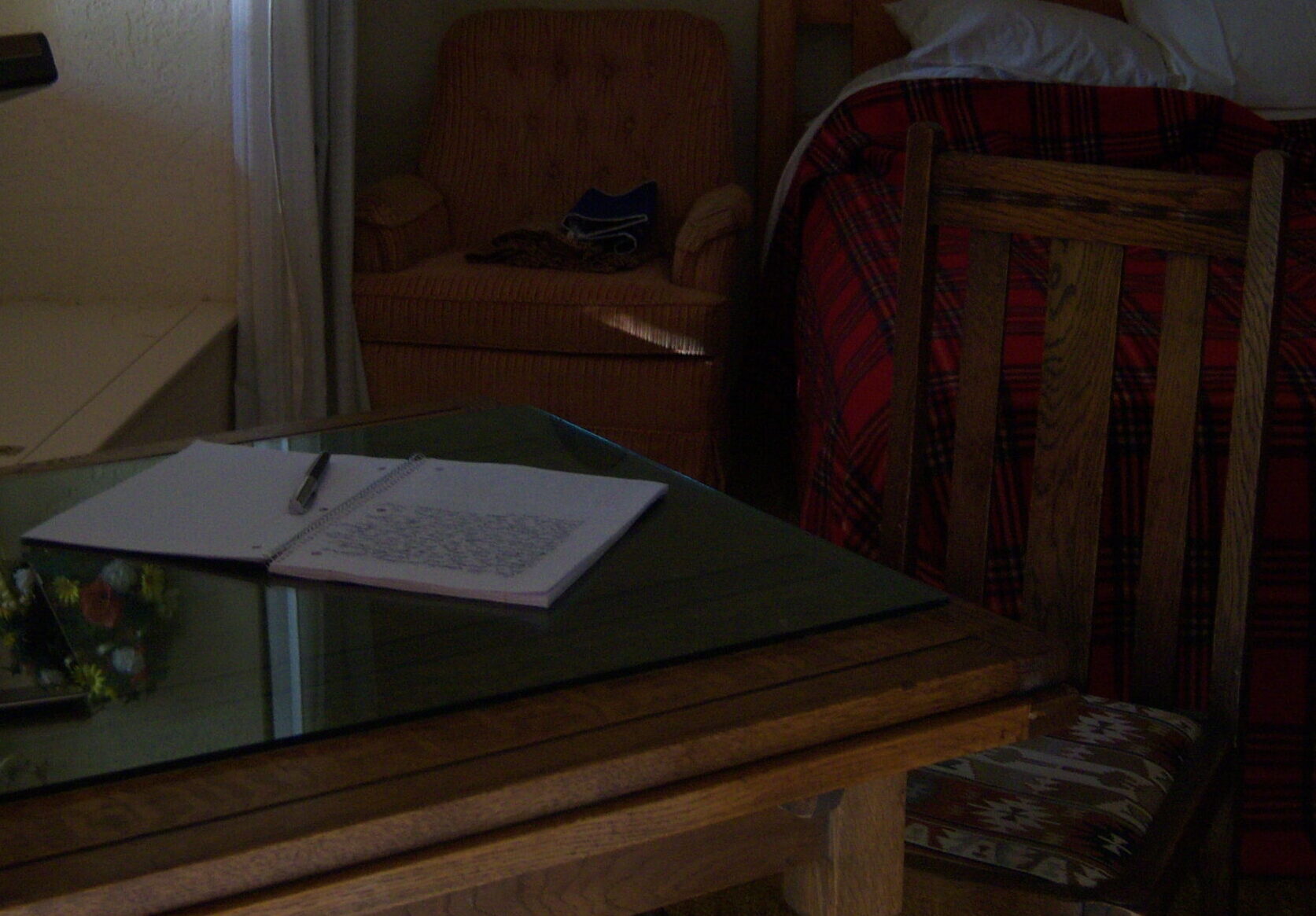 A dimly lit room with a table and open notebook, a bed with plaid bedding, an armchair, a lamp, and wall decorations.