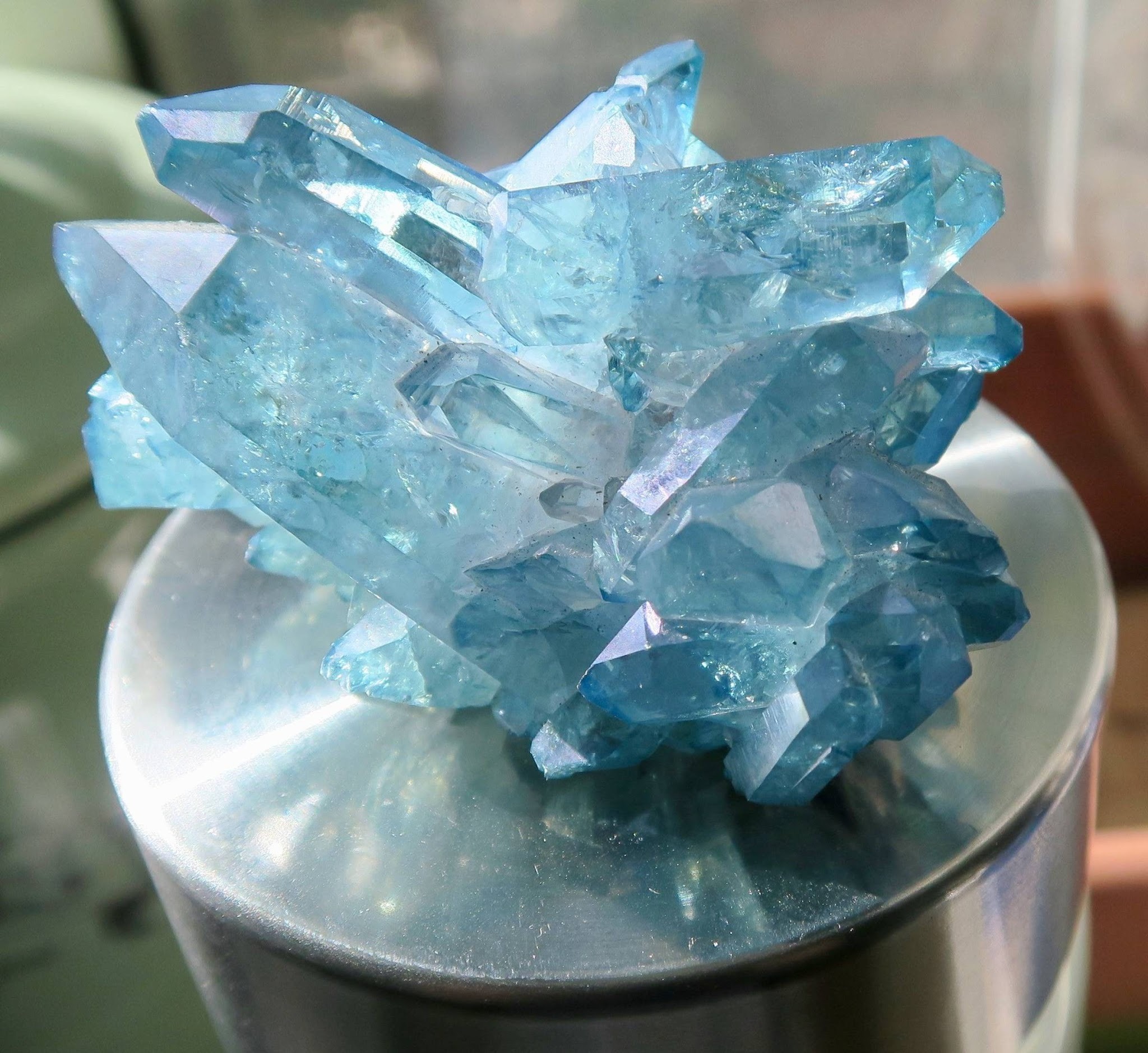 A cluster of blue crystals sits on a metallic cylindrical surface, reflecting light and showing multiple clear, angular facets.