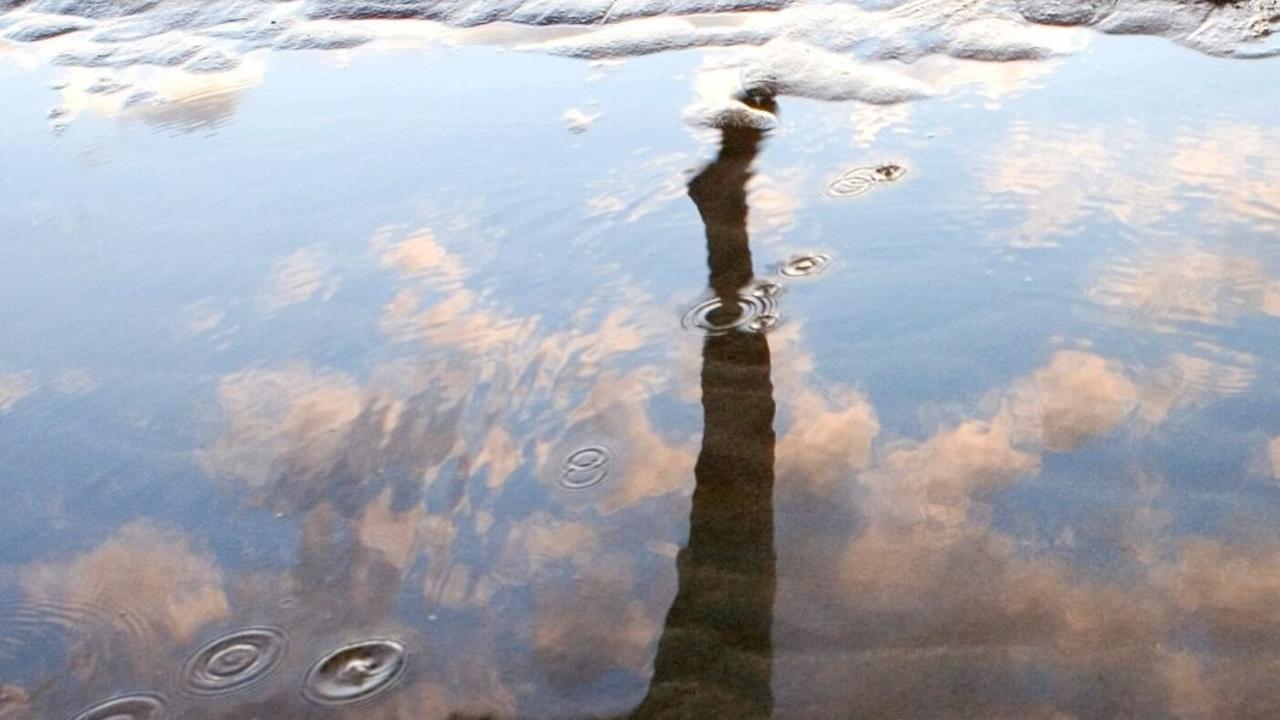 A lamppost and clouds are reflected in a puddle, with raindrops creating ripples on the water's surface.