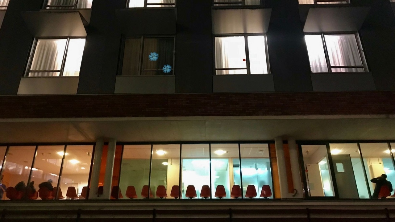 Exterior view of a building at night showing rows of orange chairs in a brightly lit interior space, with several upper windows partially open and curtains drawn.