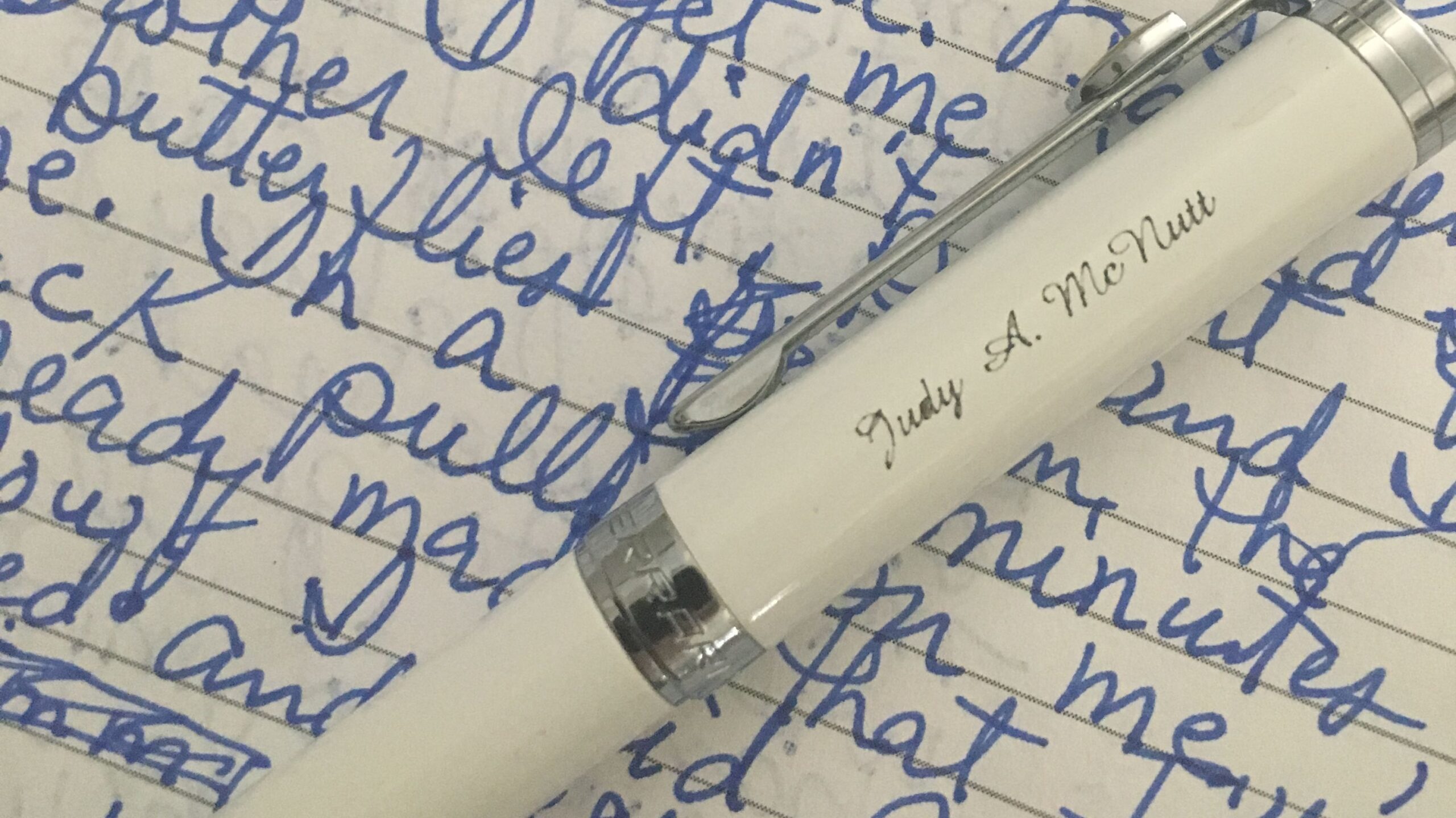 A white pen engraved with "Judy A. McNutt" rests on a page filled with blue handwritten cursive text.