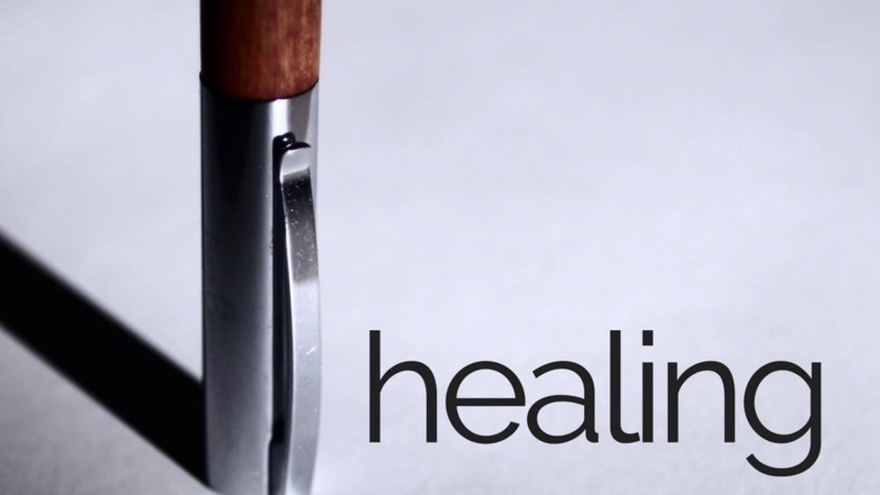 A close-up of a metallic pen casting a shadow, with the word "healing" in lowercase letters to the right.